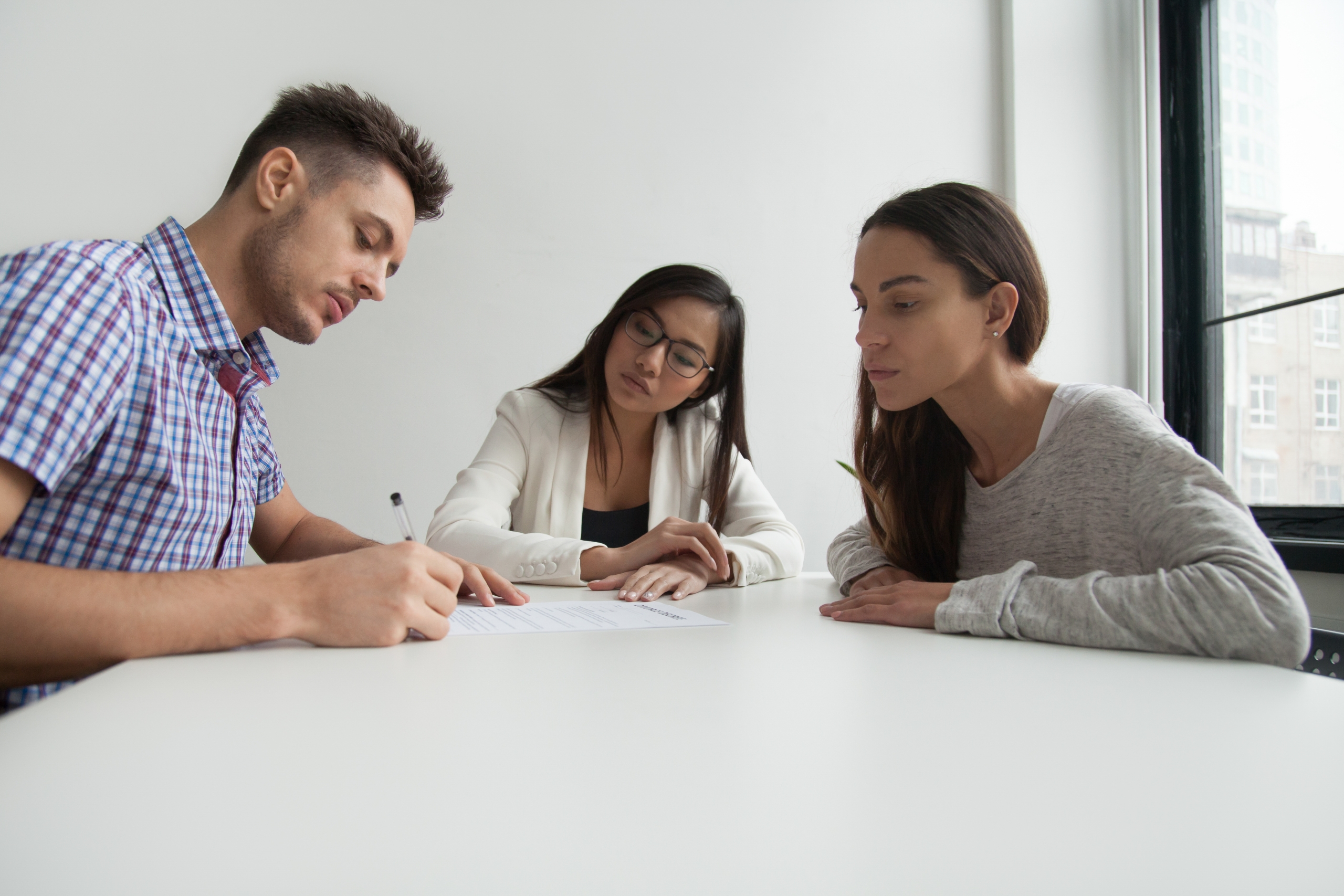 Unhappy husband signing divorce papers, concerned wife watching waiting for her turn in office of Asian lawyer observing documentation and legalizing couple break up. Concept of unsuccessful marriage